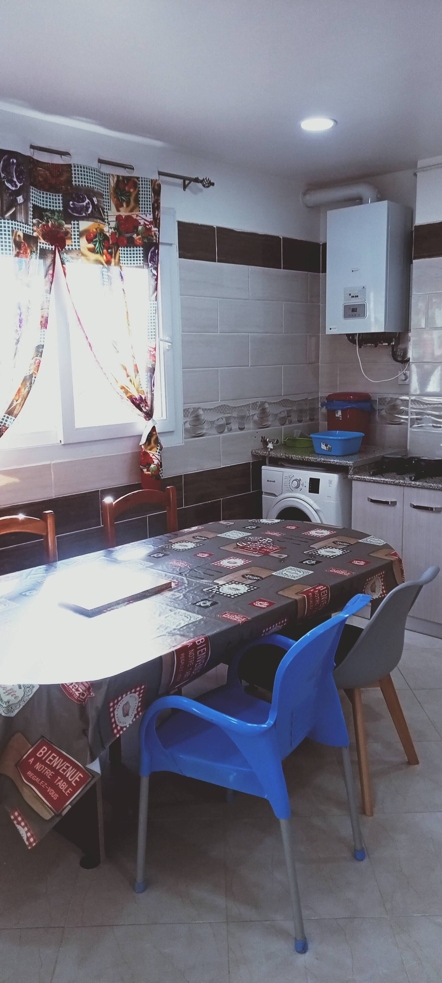 Een eethoek met een ronde tafel, bedekt met een tafelkleed met dessin, blauwe plastic stoelen en een bordje met de tekst 'Bienvenue A Notre Table'.