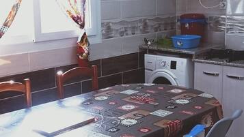 Een eethoek met een ronde tafel, bedekt met een tafelkleed met dessin, blauwe plastic stoelen en een bordje met de tekst 'Bienvenue A Notre Table'.