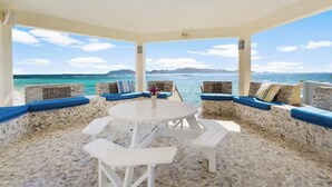 Un espacio de estar al aire libre con techo, mesas y bancos, paredes de piedra y vista al mar.