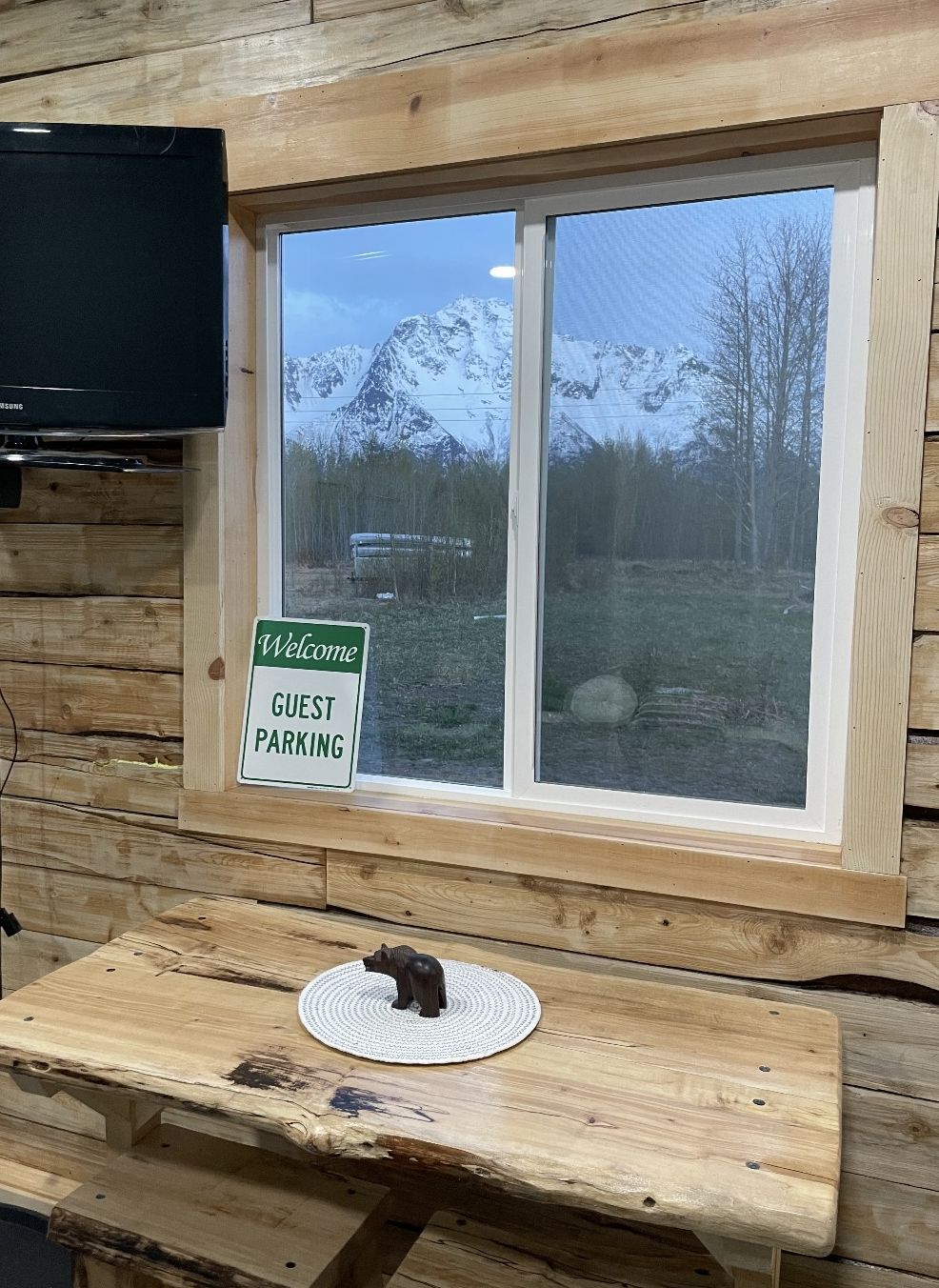 A wooden cabin with a 'Welcome Guest Parking' sign, a television mounted on the wall, and a wooden table with a placemat and a small figurine.