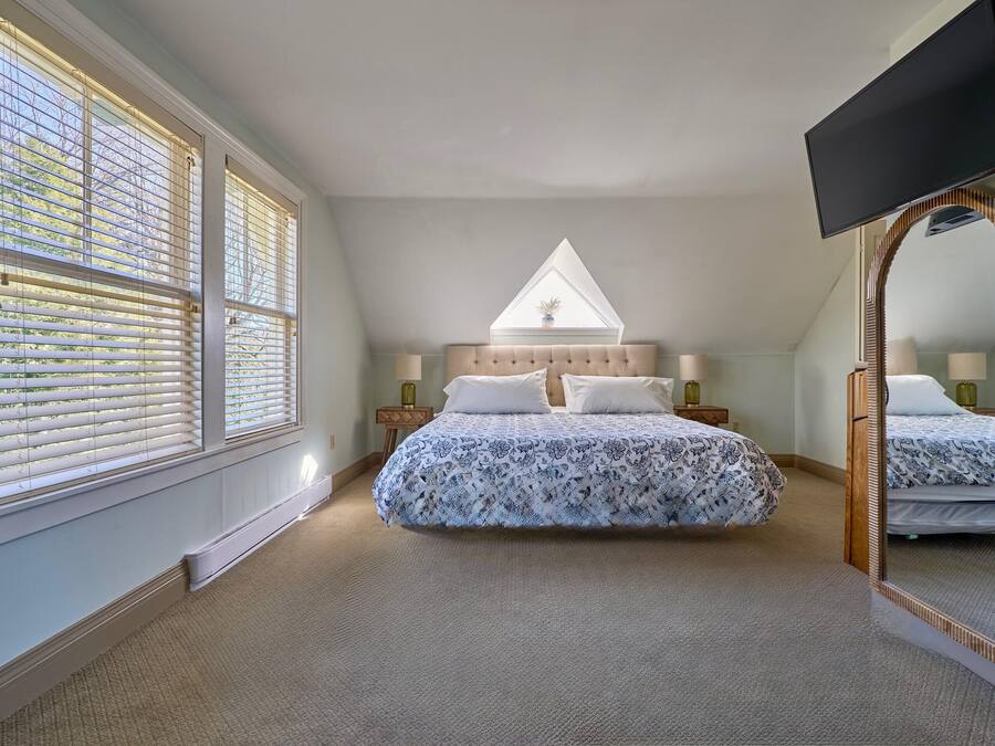 A bedroom with a large bed, a window with blinds, a ceiling fan, and a flat-screen TV mounted on the wall.