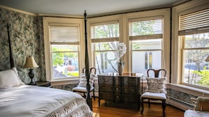 Un dormitorio con una cama grande, un aparador, una silla y una mesa. Hay tres ventanas con persianas, y una lámpara está sobre una mesita de noche.