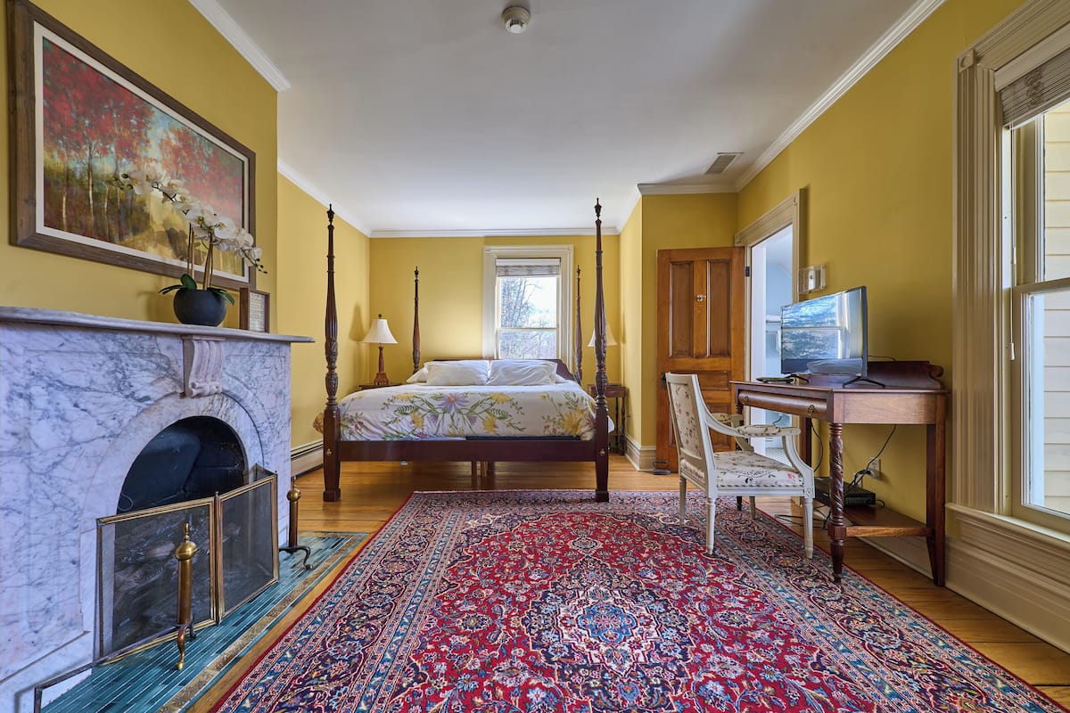 Un dormitorio con cama, chimenea, escritorio y ventana grande.