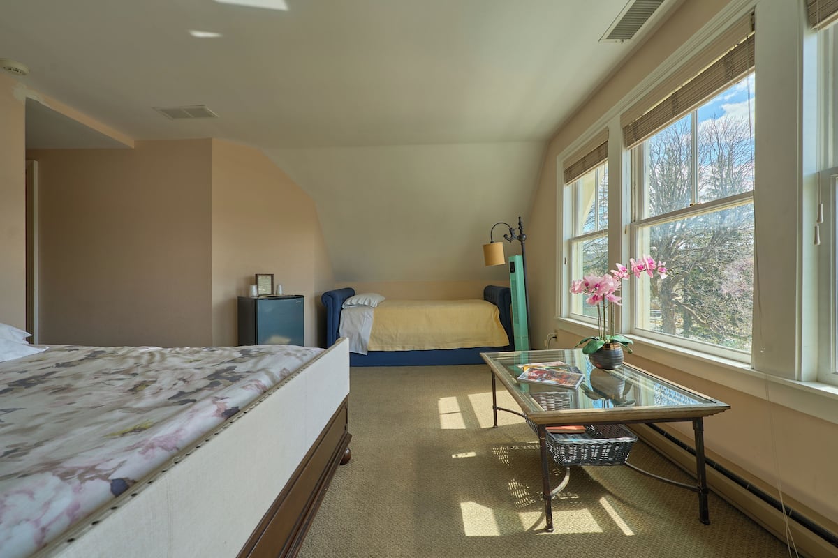 Un dormitorio con cama, escritorio, silla, ventana con persianas y vista a árboles.