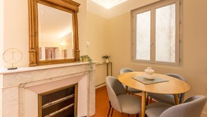 A dining area with a round table, grey chairs, a wooden side table, a mirror, and a window with blinds.