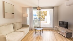 Ein modernes Wohnzimmer mit einer Couch, einem kleinen runden Tisch, einem Fernseher auf einem Schrank und einer großen Schiebeglas-Tür, die auf einen Balkon im Freien führt.