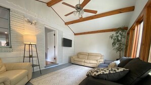 Een ruime woonkamer met een hoge plafond, een plafondventilator, een beige bank, een donkere bank, een getextureerd vloerkleed en een potplant.