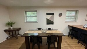 Un comedor con una mesa rectangular y cuatro sillas, una mesita auxiliar con una planta y dos ventanas con persianas.