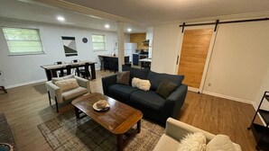 Una sala de estar moderna con un sofá, sillones y una mesa de centro. En el fondo, hay una zona de comedor con una mesa y sillas.