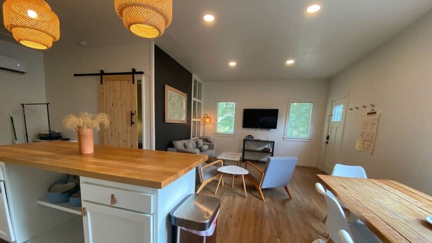 Une cuisine moderne avec un îlot central en bois, des meubles blancs et une poubelle. On y trouve également un coin repas avec une table et des chaises, ainsi qu’un téléviseur fixé au mur.