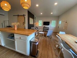Een moderne keuken met een houten kookeiland, witte kasten en een afvalbak. Er is een eethoek met een tafel en stoelen, en een televisie aan de muur bevestigd.