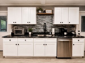 A modern kitchen with white cabinets, a black countertop, a built-in oven, a coffee maker, and a dishwasher. - Dollywood Cabin - Luxury with Private Arcades (Sevier County)
