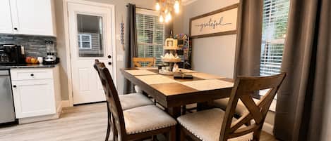A dining area with a wooden table, cushioned chairs, a chandelier, and a wall with a 'grateful' sign.