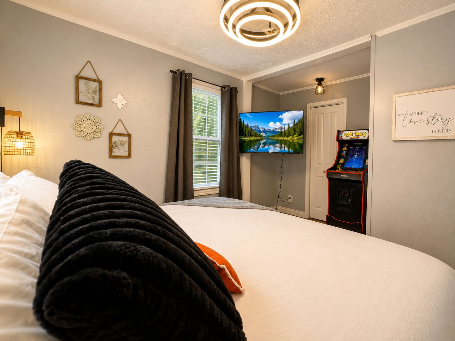Una habitación de hotel con cama, televisión, máquina arcade de Pac-Man y ventana con cortinas.