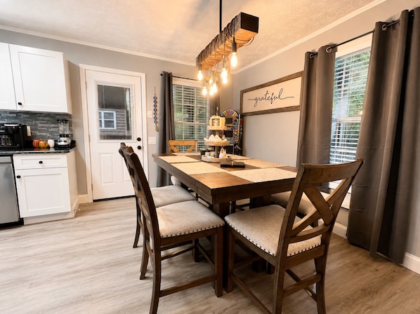 A dining area with a wooden table, cushioned chairs, a chandelier, and a wall with a 'grateful' sign.