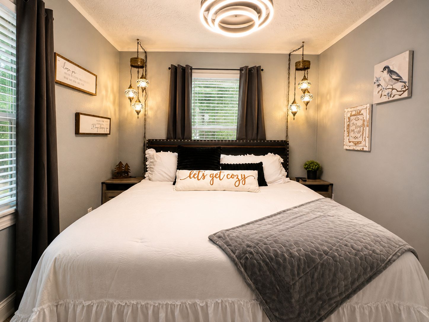 A bedroom with a large bed, two bedside tables, a nightstand, a wall-mounted light fixture, and a window with curtains.