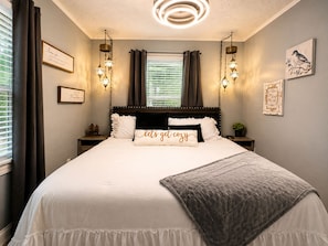 A bedroom with a large bed, two bedside tables, a nightstand, a wall-mounted light fixture, and a window with curtains.