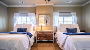 Quarto de hotel com duas camas, uma mesinha de cabeceira de madeira, uma luminária e um quadro na parede.