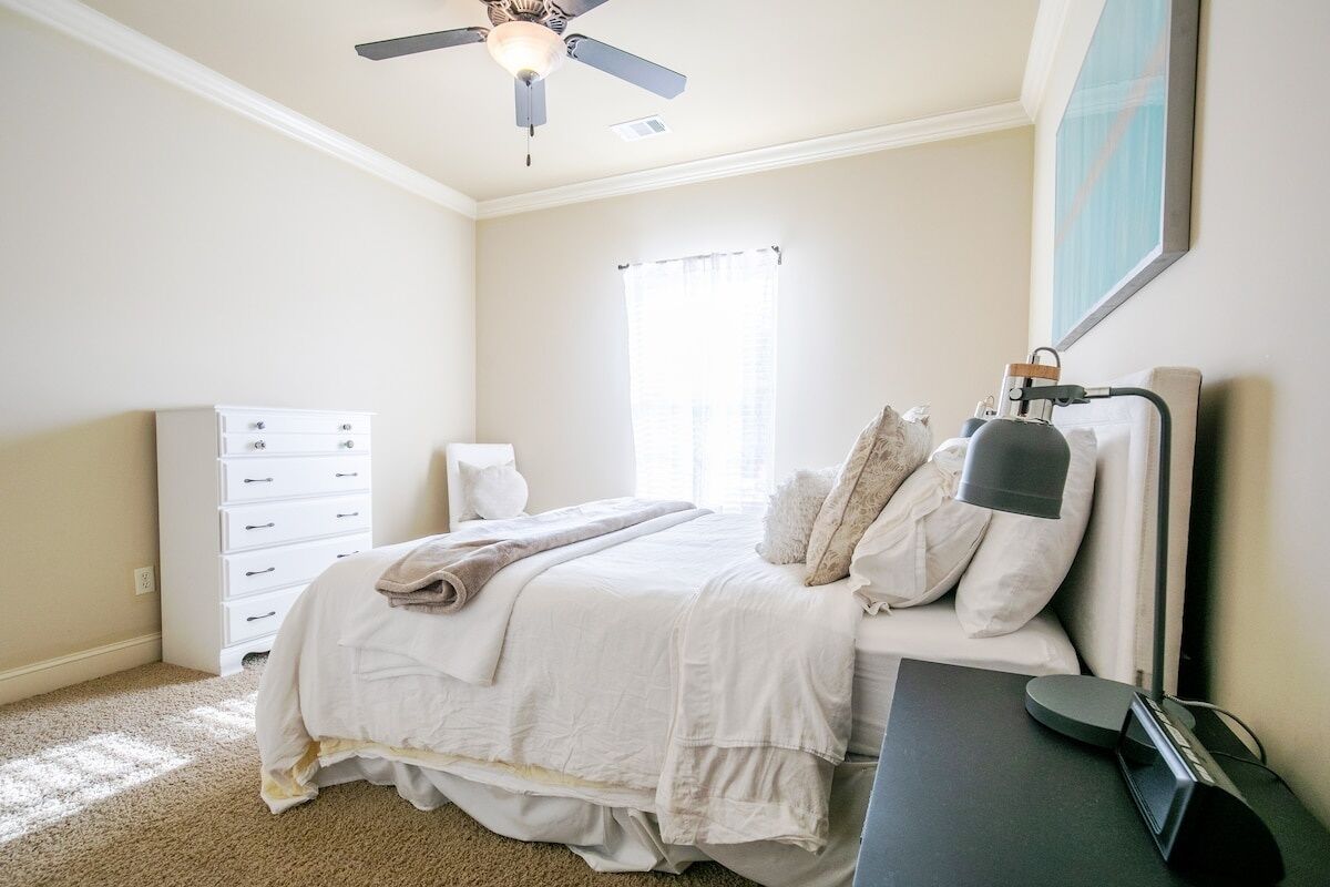 A bedroom with a bed, a dresser, a desk with a lamp, a ceiling fan, and a window with curtains.