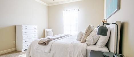 A bedroom with a bed, a dresser, a desk with a lamp, a ceiling fan, and a window with curtains.