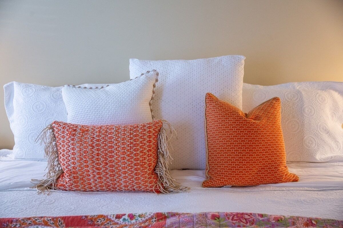 Una cama con almohadas blancas y dos almohadas anaranjadas con estampado.