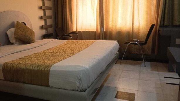 A neatly made bed with a patterned bedspread, two pillows, and a chair in a room with a tiled floor and curtains. - Grande Sita Hotel (Udaipur)
