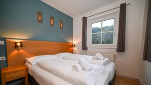 Un letto matrimoniale con lenzuola e asciugamani bianchi, un comodino in legno con cassetto, una lampada a parete e una finestra con tende.