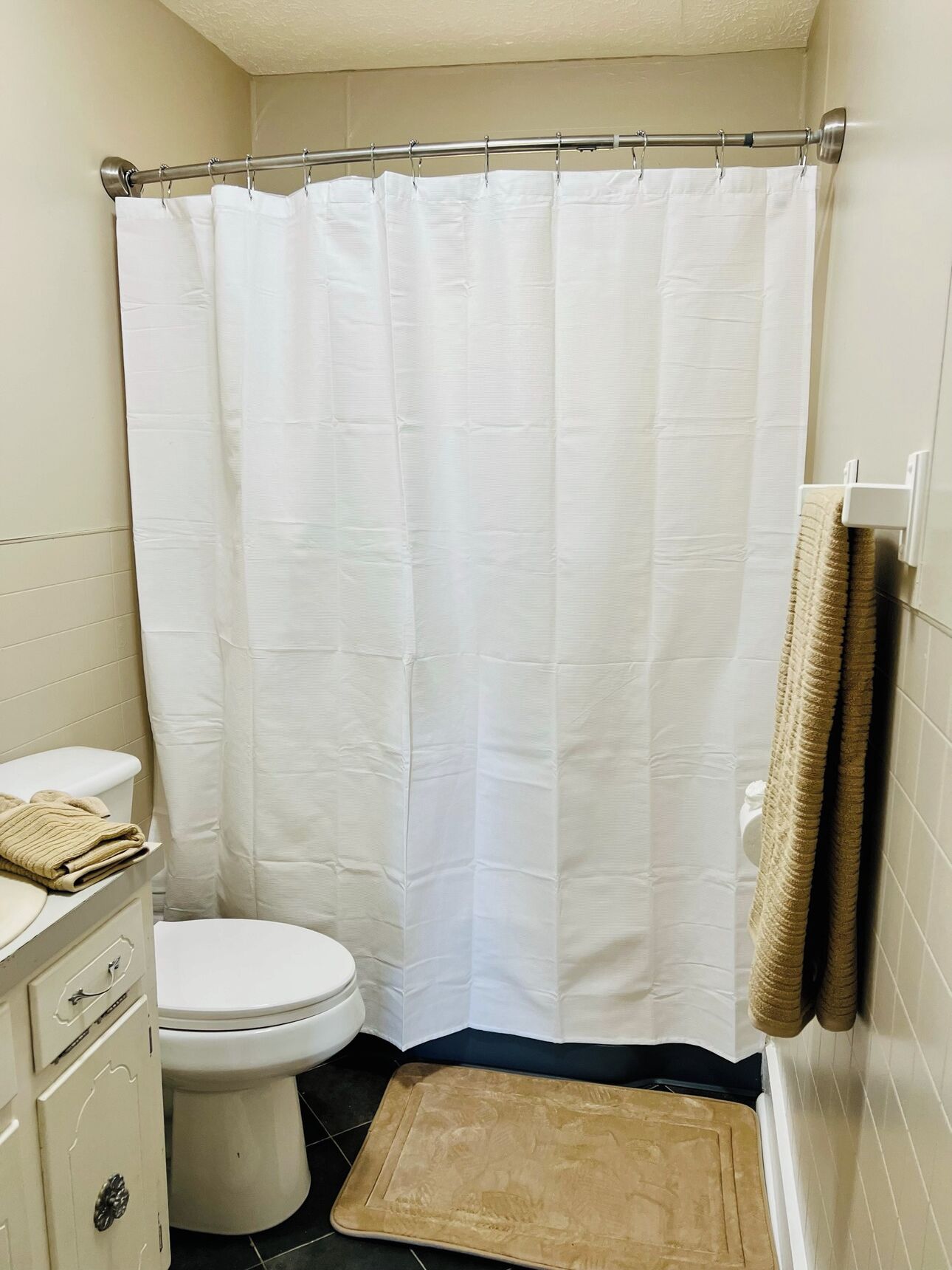 A small bathroom with a white shower curtain, a toilet, a beige mat, and a towel rack with towels.