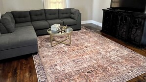 A living room with a sectional sofa, a glass coffee table, a patterned rug, a television, and a dark wooden cabinet.