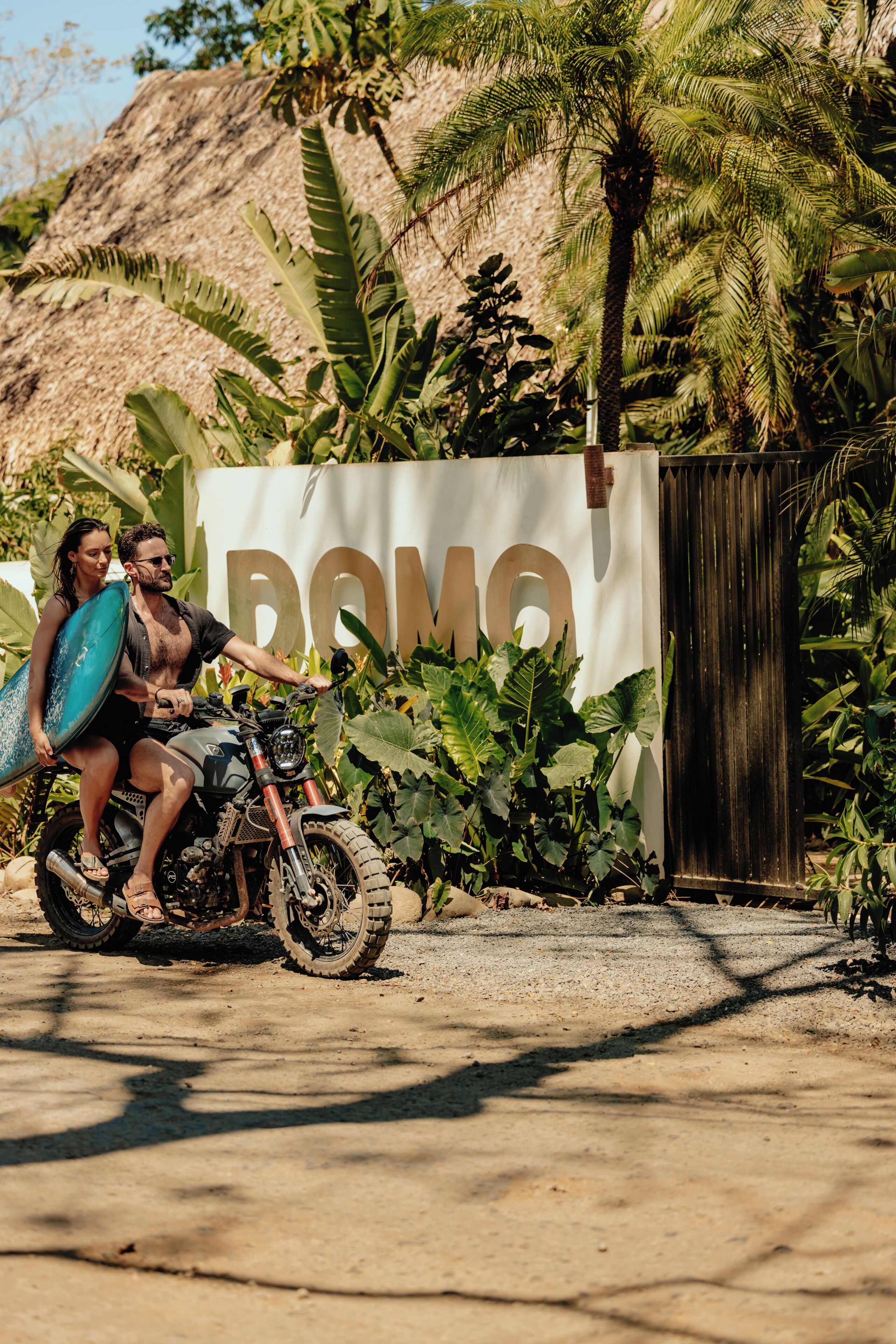 Een motor met twee passagiers, een bord met de tekst 'ROMA' en tropische begroeiing.