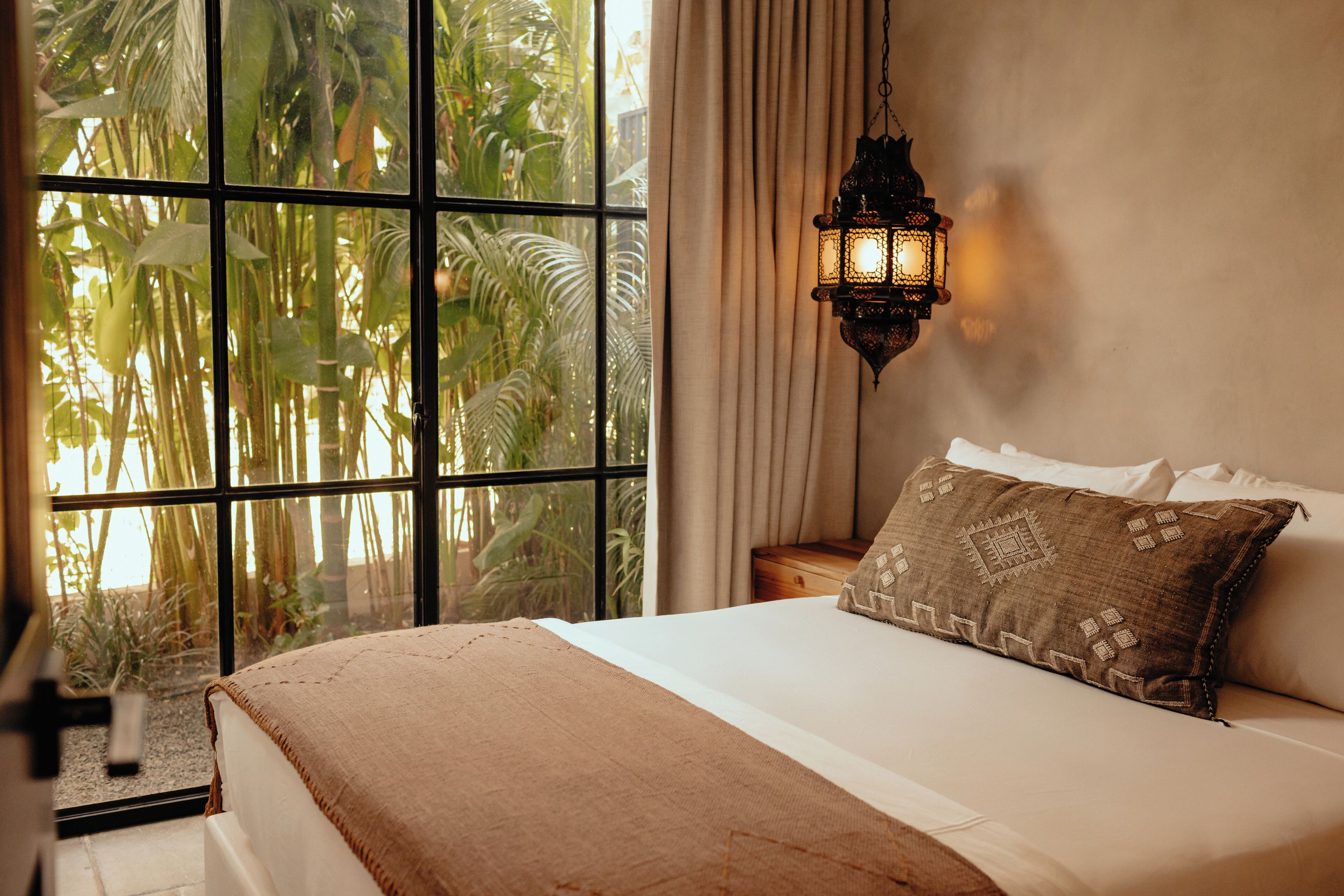 A bedroom with a large window, a bed with a brown blanket, a patterned pillow, and a hanging lantern.