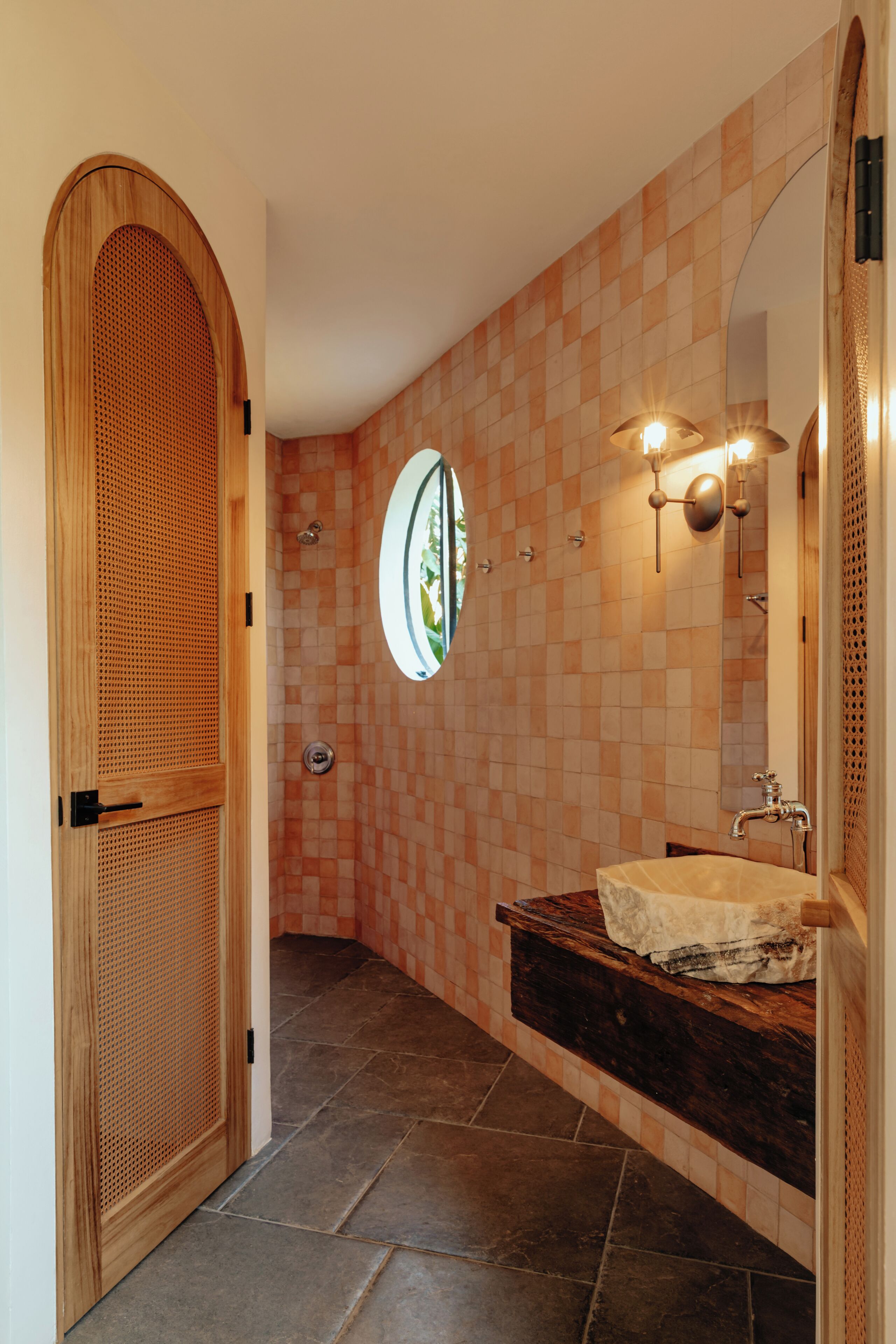 Ein Badezimmer mit Holztür, rundem Spiegel, Waschbecken mit Natursteinplatte und gefliesten Wänden.