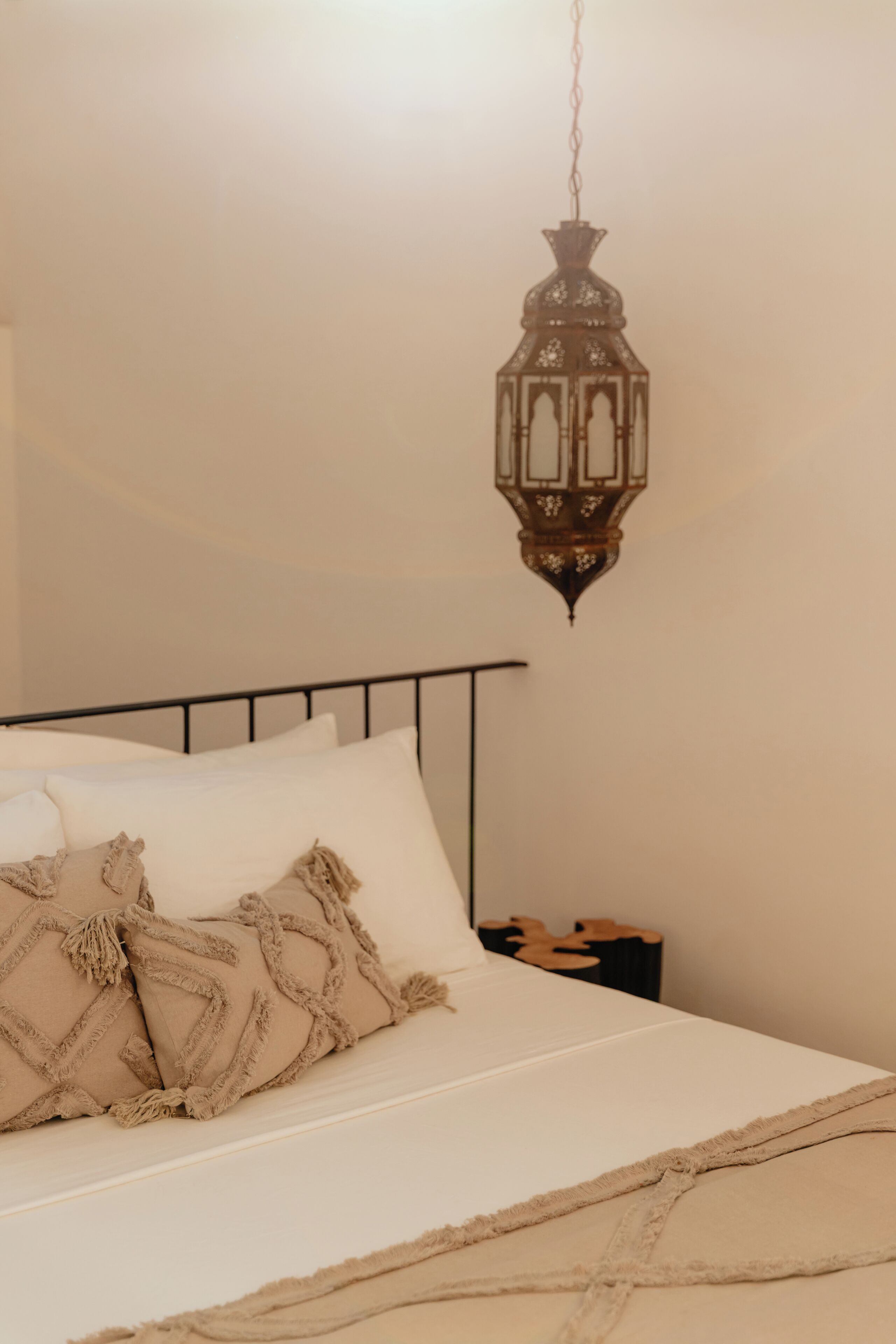 Una cama con ropa de cama blanca y almohadas color beige, un farol colgante y un cabecero de metal negro.