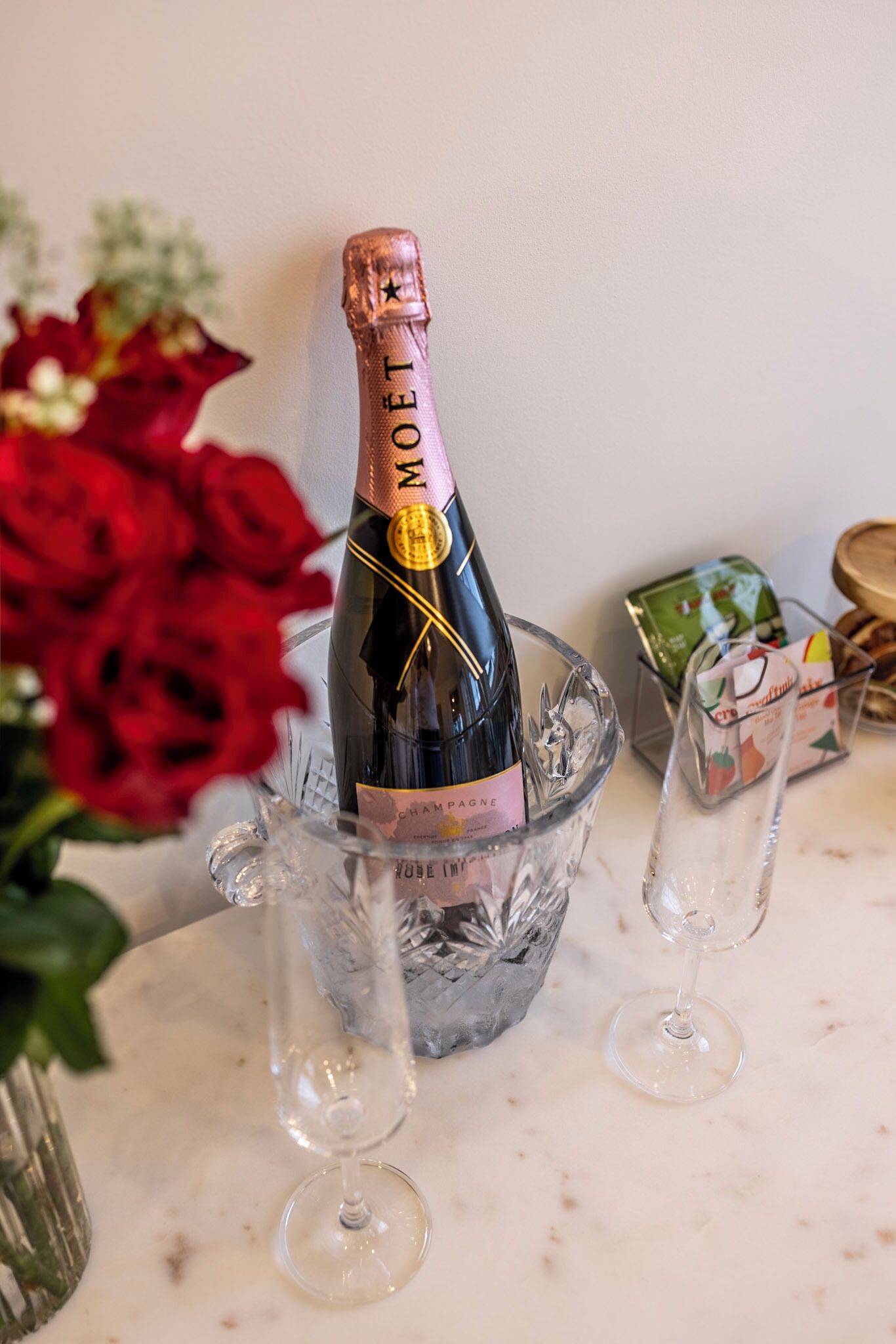 A bottle of champagne in a glass ice bucket with two empty glasses beside it, surrounded by red roses and a small gift box.