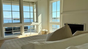 A bedroom with a large window overlooking a body of water, a fireplace, and a bed with pillows.