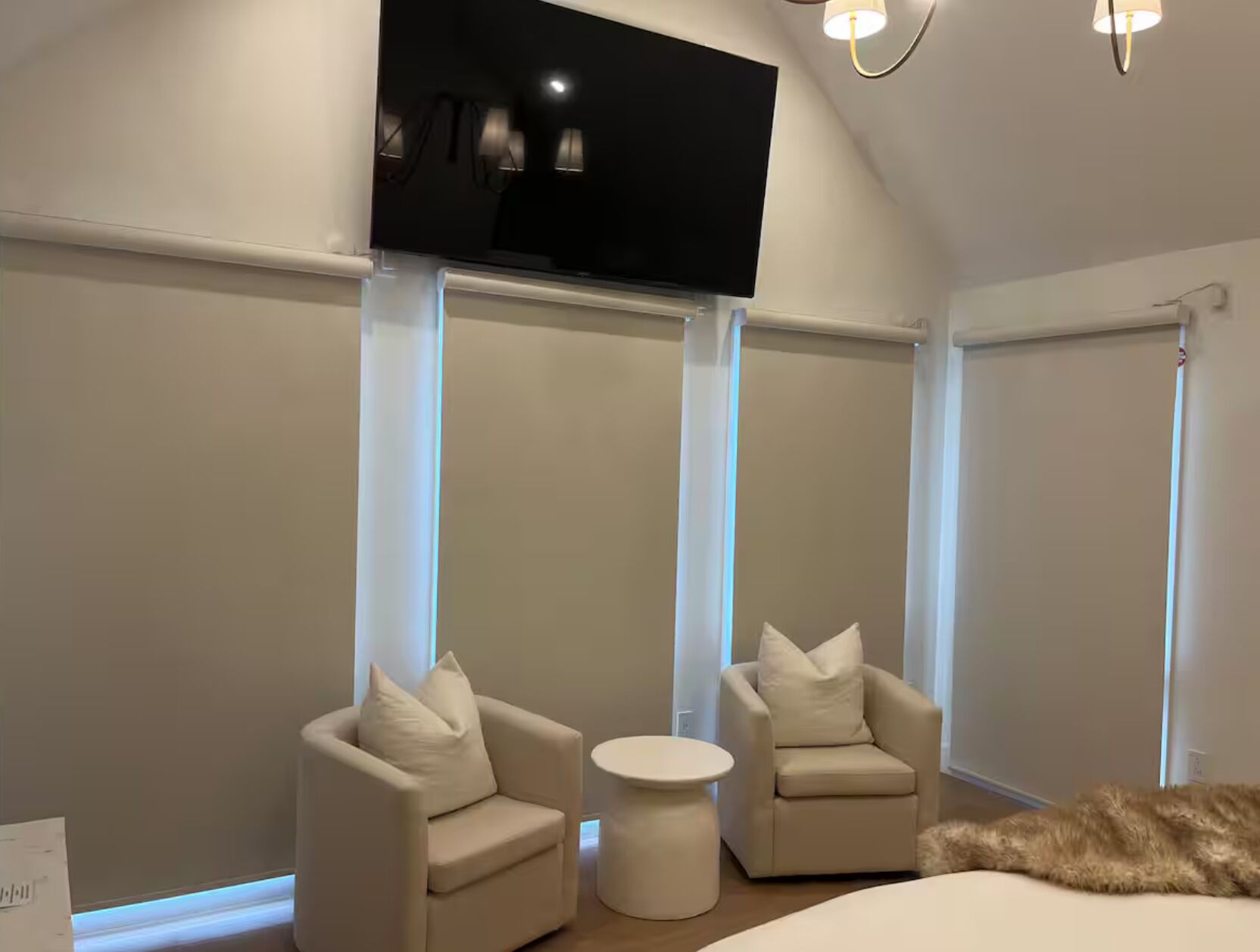 A modern bedroom with a large bed, two armchairs, a small round table, and a TV mounted on the wall.