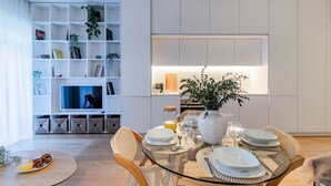 A modern dining area with a glass table set for four, surrounded by wicker chairs. A bookshelf filled with books and decorative items is visible in the background.
