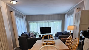A dining area with a wooden table and chairs, a television in the background, and a kitchen area with cabinets and a sink. - Thoughtfully Renovated 2BR Retreat|10 Miles to South Haven & Lake Michigan (Bangor)