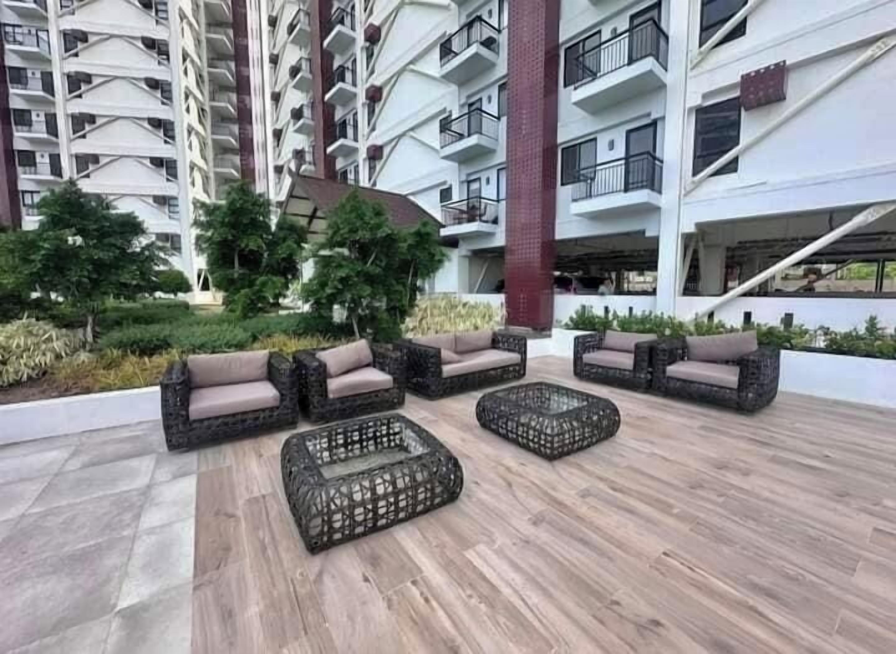 Un espace extérieur moderne aménagé avec des meubles en osier et un plancher en bois, entouré de verdure et d’un immeuble de grande hauteur doté de balcons.