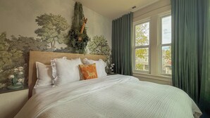 Een slaapkamer met een groot bed, een hoofdbord met muurschildering, een raam met gordijnen, en een nachtkastje met lamp.