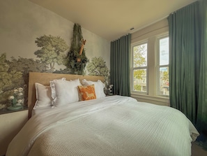 Une chambre avec un grand lit, une tête de lit ornée d’une fresque murale, une fenêtre avec des rideaux et une table de chevet avec une lampe.