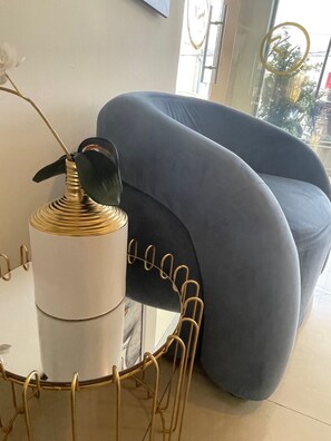 Una sala de estar moderna con un sofá gris, un objeto cilíndrico blanco y un objeto negro encima, y una mesa con estructura de alambre dorada.