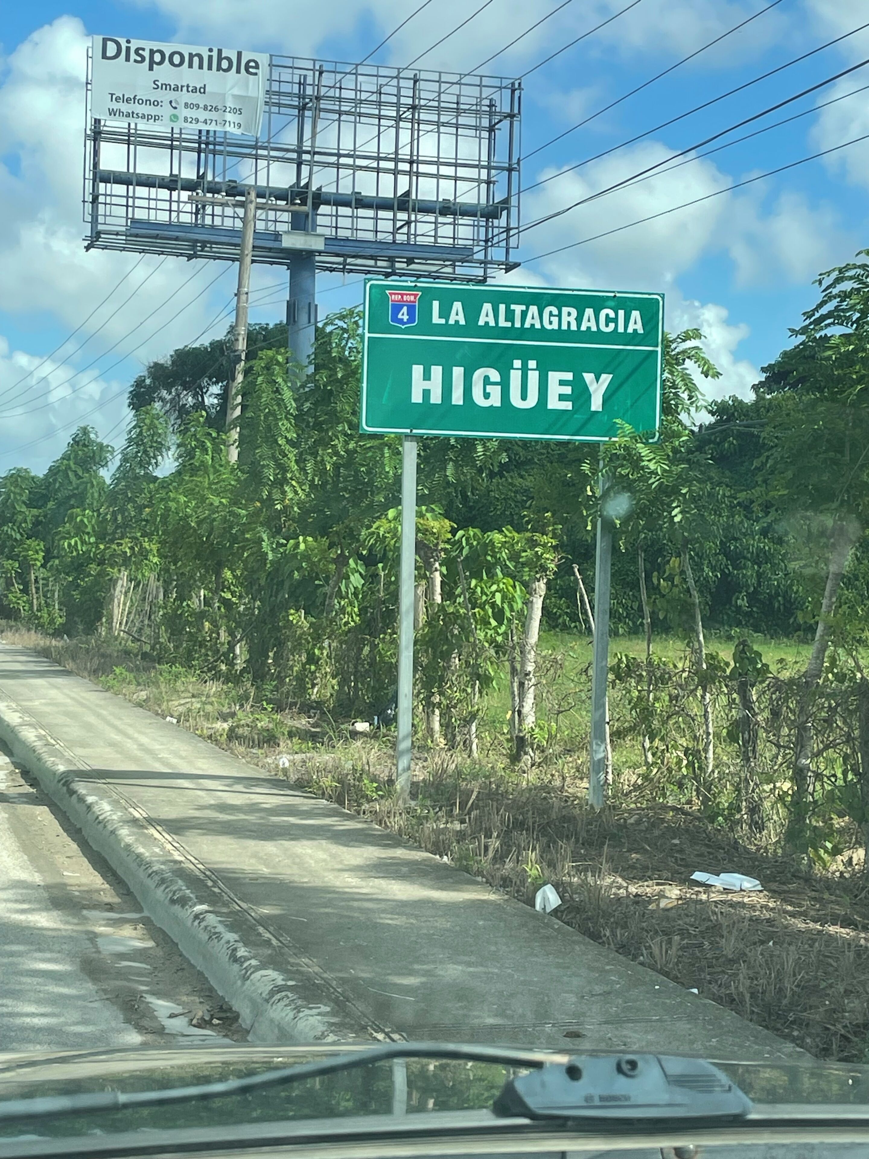 Señal de tráfico que indica 'LA ALTAGRACIA HIGUEY' con un cartel publicitario cercano que anuncia 'Disponible'.
