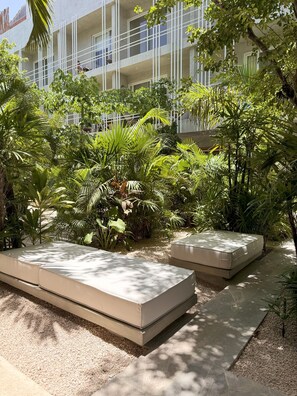 A courtyard with two beige outdoor seating areas surrounded by lush greenery and a building with a modern design in the background.