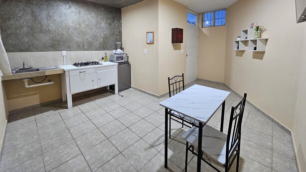Cocina compacta con estufa a gas, pileta y heladera pequeña. Cuenta con mesa y sillas, y una ventana con cortinas azules.