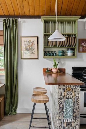 A kitchen with a wooden countertop, green curtains, a hanging pendant light, and a patterned backsplash.