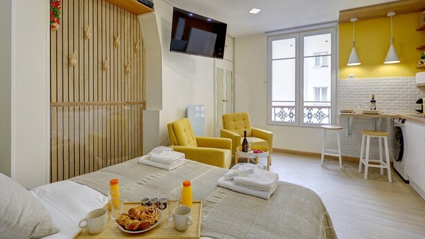 A modern hotel room with a bed, two yellow armchairs, a TV, a balcony view, and a kitchenette.
