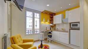A compact kitchen with white cabinets, a microwave, and a sink. There is a yellow chair and a small round table with a bottle and glasses.