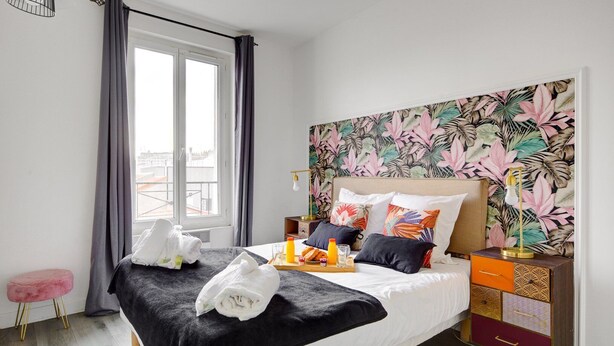 A modern hotel room with a large bed, a tropical leaf-patterned headboard, a bedside table with a lamp, and a small stool. - TBS 140 - Elegantly decorated apartment (Paris)
