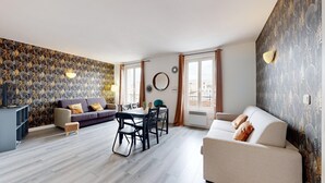 A modern living room with a sofa, dining table, and chairs. There is a TV on a stand, a small table with decorative items, and a window with curtains. - TBS 140 - Elegantly decorated apartment (Paris)